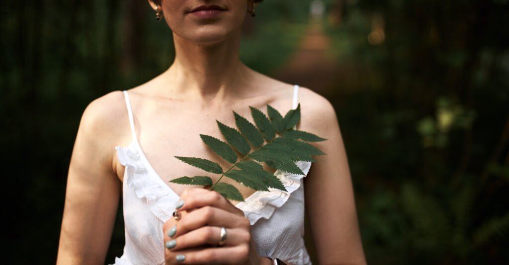 cropped shot of beautiful tender young bride in romantic white dress posing against green forest background, holding fern leaf at her chest. unrecognizable female relaxing outdoors among plants cropped shot of beautiful tender young bride in romantic white dress posing against green forest background, holding fern leaf at her chest. unrecognizable female relaxing outdoors among plants